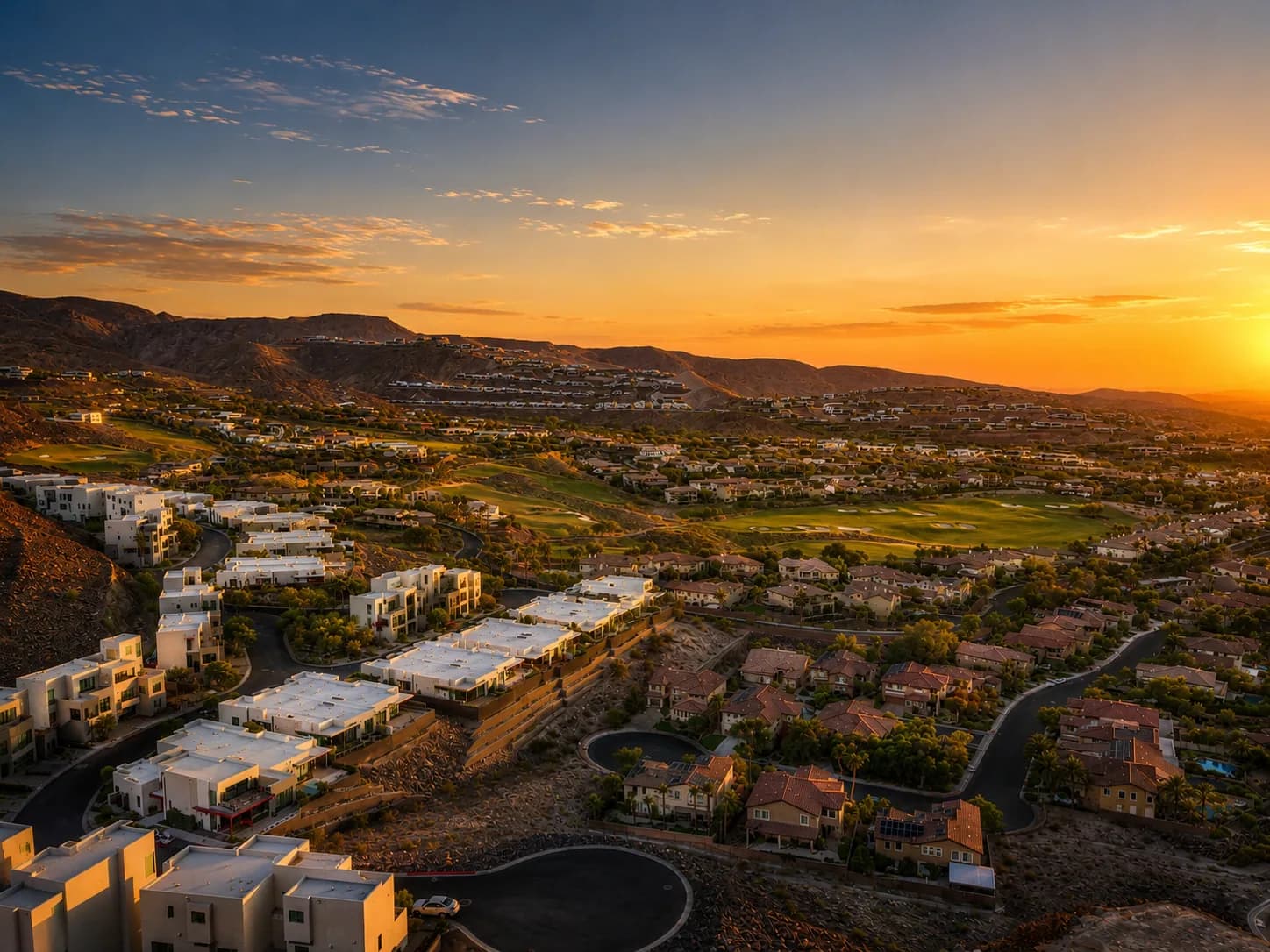 Aerial view of a Las Vegas hillside neighborhood at golden hour — homes and a green golf course below the desert mountains